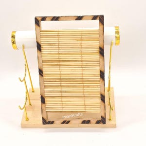 May include: A jewellery display stand with a light wood base and a bamboo-style frame. The frame has dark brown accents and holds a bamboo mat. White cylindrical holders with gold accents are on each side. The stand has gold hooks.