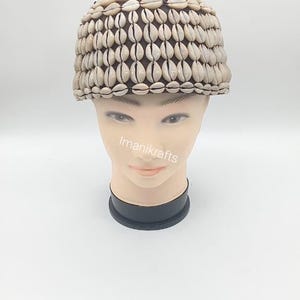 Peut inclure: Un chapeau beige et marron composé de petits coquillages. Le chapeau est posé sur une tête de mannequin avec le texte "Imanikrafts" sur le devant. Le chapeau est de forme arrondie et est probablement fait à la main.