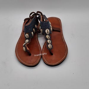 May include: A pair of brown leather sandals with a thong-style toe strap. The sandals feature a blue beaded strap adorned with white seashells. The sandals have a brown ankle strap and are handmade.