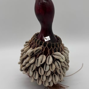May include: A handcrafted percussion instrument, likely a gourd shaker, featuring a deep burgundy body and a skirt of white cowrie shells. The shells are attached to a woven net, and a small tag is visible. A brown string is attached to the top.