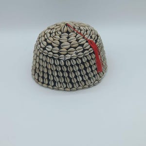 Op de afbeelding: Een handgemaakte, koepelvormige hoed bedekt met kleine, beige schelpen. De hoed heeft een zwart interieur en een decoratieve rode kwast. Het item is waarschijnlijk een uniek modeaccessoire of decoratief stuk.