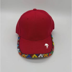 Puede incluir: Gorra de béisbol roja con visera curva. La visera está decorada con coloridos patrones de cuentas. Un pequeño logotipo de elefante blanco está bordado en la parte delantera. La gorra está hecha de tela roja lisa.