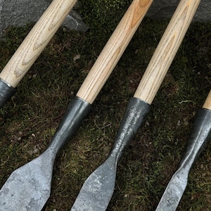 May include: Close-up of four garden tools with wooden handles and metal blades. The handles are light-coloured wood, and the blades are a dark, textured metal. The tools are arranged on a bed of green moss.