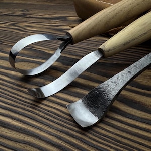 May include: Close-up of three wood carving tools with wooden handles and curved steel blades. The tools are arranged on a dark wood surface, showcasing their craftsmanship and design. The blades have a polished finish.