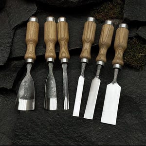 Op de afbeelding: Een set van zes houtsnijbeitels met houten handvatten en metalen bladen. De beitels zijn gerangschikt op een donker, getextureerd oppervlak, waarbij hun verschillende blad vormen en maten worden getoond. De handvatten hebben een natuurlijke houtnerf afwerking.