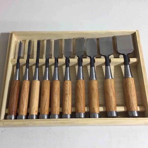 Puede incluir: Un juego de nueve cinceles para tallar madera con hojas de metal y mangos de madera, dispuestos en una bandeja de madera clara. Los cinceles tienen diferentes formas y tamaños de hoja, adecuados para proyectos de carpintería.