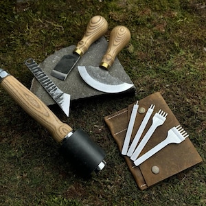 May include: A selection of leathercraft tools, including a mallet, knives, and punches, arranged on a mossy surface. The tools have wooden handles and metal blades, and are displayed with a brown leather case.
