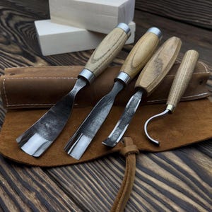 May include: A set of four wood carving tools with wooden handles and metal blades, displayed on a brown leather tool roll. The tools include a curved gouge, a straight gouge, and a hook knife. Two small wooden blocks are in the background.