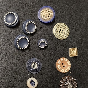 May include: A selection of vintage buttons in diverse shapes and colours. The buttons feature navy blue, cream, brown, and beige hues. Some buttons display intricate designs, including floral patterns and geometric shapes. The buttons are arranged on a dark surface.