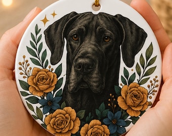 May include: A round white ceramic ornament featuring a detailed illustration of a black dog's face centered among golden-yellow roses, blue flowers, and green leaves. The ornament has a small hole at the top with a thin gold hanging cord.