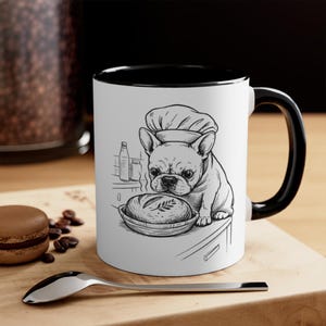May include: White ceramic mug with a black interior and handle. The mug features a black and white illustration of a French Bulldog wearing a chef's hat, looking at a loaf of bread. The mug is on a wooden surface.