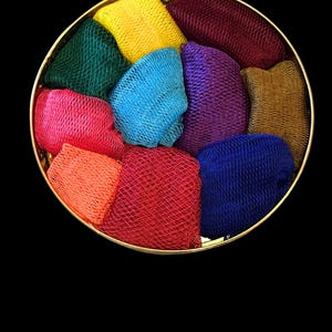 May include: A round gold-colored container filled with ten small, folded mesh scrubbers in various vibrant colors including yellow, dark red, green, bright blue, purple, pink, orange, red, dark blue, and brown, arranged in a circular pattern.