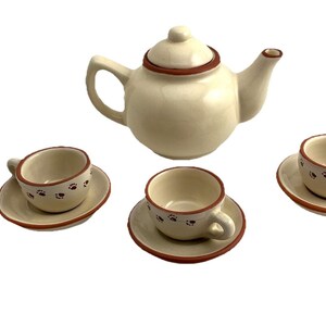 May include: Cream-colored ceramic tea set with a teapot and three teacups on saucers. The set has brown paw print details and brown trim. A charming addition to any tea party.