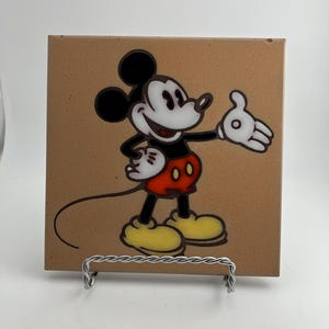 May include: A square ceramic tile featuring a cartoon image of Mickey Mouse. Mickey is depicted in his classic outfit with black shorts, yellow shoes, and white gloves. The tile has a brown background and is displayed on a silver wire stand.