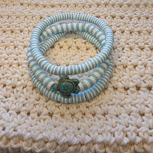 Puede incluir: Tres pulseras con cuentas a rayas azules y blancas, aseguradas con un cierre de tortuga turquesa. Las pulseras están sobre una superficie texturizada de color crema.