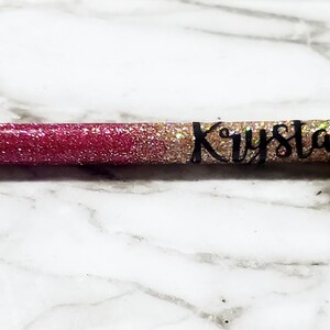 Może przedstawiać: Błyszczący długopis z czarną końcówką i srebrnym metalowym końcem. Długopis jest ozdobiony różowym i złotym brokatem oraz imieniem "Krystal" czarnym pismem. Długopis leży na białej i szarej marmurowej powierzchni.