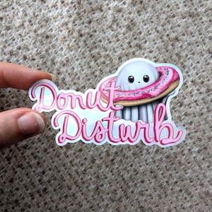 Peut inclure: Un autocollant découpé représentant une créature blanche, ronde et tentaculaire portant un beignet rose glacé avec des vermicelles colorés en guise d'anneau. Le texte Donut Disturb est écrit en police cursive rose sur le devant.