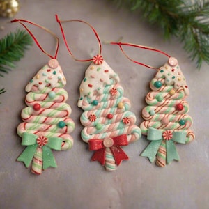 May include: Three Christmas tree ornaments. Each tree is made of candy canes in red, white, and green, topped with white icing and sprinkles. A red ribbon is attached for hanging, and a green bow is at the base.