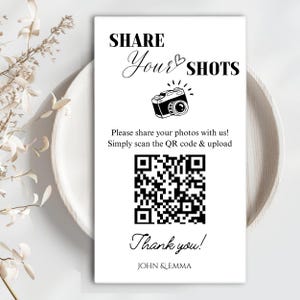 May include: A rectangular white sign with black text and graphics. The text reads SHARE Your SHOTS, Please share your photos with us! Simply scan the QR code & upload, Thank you!, and JOHN & EMMA. A camera icon and a large QR code are centered.