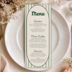 Könnte beinhalten: Eine vertikale, rechteckige Menükarte mit hellbeiger Mitte und salbeigrünen Streifenrändern. Das Design zeigt einen dunkelgrünen, bogenförmigen Rahmen mit dem Text Menu, Starters, Main Dishes und Desserts sowie detaillierten Speisebeschreibungen.