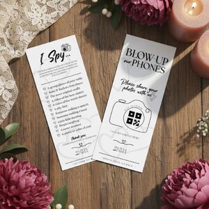 May include: Two white cards with black text on a wooden surface. One card says "I Spy..." with a list of prompts. The other says "BLOW UP our PHONES" with a camera graphic and QR code. Pink flowers and candles surround the cards.