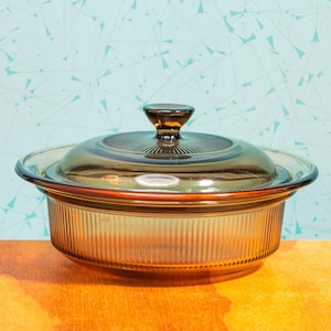 Puede incluir: Una fuente de cristal ámbar con tapa. La fuente tiene forma redonda con lados acanalados verticales y una tapa a juego con un asa de pomo. La fuente está sobre una superficie de madera con un fondo azul claro.