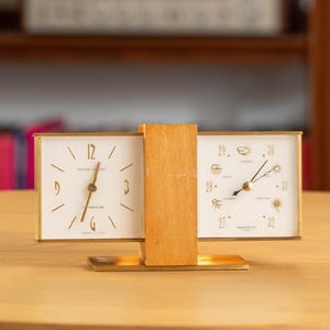 Puede incluir: Un reloj y una estación meteorológica vintage. El reloj tiene una esfera blanca con detalles dorados y un marco dorado. La estación meteorológica tiene una esfera blanca con símbolos meteorológicos. El reloj y la estación meteorológica están conectados por un bloque de madera.