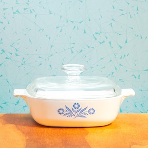 May include: A white casserole dish with a clear glass lid and a blue floral design. The dish has two handles and sits on a wooden surface. The background is a light blue wall with a pattern of blue lines and shapes.