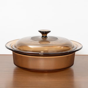 May include: A round, amber-coloured glass casserole dish with a matching lid. The base features vertical ribbing along the exterior, while the lid has a central knob handle and a circular, sunburst-patterned texture surrounding the knob.