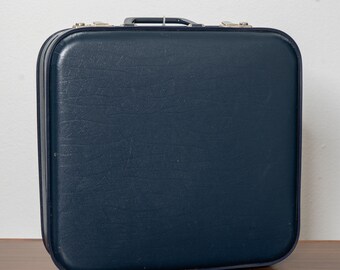 May include: A square, dark blue suitcase with a textured, leather-like finish. The suitcase features a dark blue top handle and silver-toned metal latches on the upper edge. The corners are rounded, and the overall design is simple and structured.