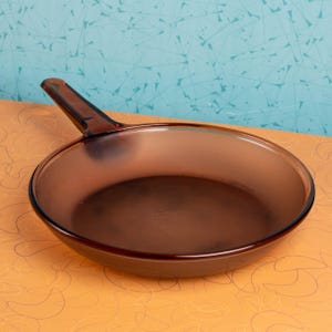 Könnte beinhalten: Eine braune Glasbratpfanne mit Griff. Die Pfanne ist rund und hat ein leicht mattiertes Aussehen. Der Griff ist an der Seite der Pfanne befestigt und ebenfalls braun. Die Pfanne steht auf einer orangefarbenen Oberfläche.