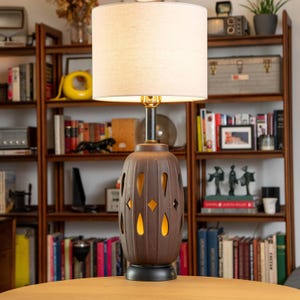 May include: A brown ceramic table lamp with a cream-colored lampshade. The lamp base has a unique design with cut-out shapes that allow light to shine through. The lamp is on a wooden table in front of a bookshelf.