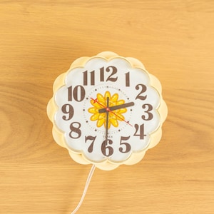 May include: A vintage, cream-colored wall clock with a flower-shaped face. The clock features a white face with black numerals and a yellow flower design in the center. The clock hands are black and red. The clock is approximately 8 inches in diameter.