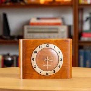 Pode incluir: Um relógio de madeira vintage com um mostrador castanho claro e um mostrador circular. O mostrador do relógio tem algarismos e ponteiros pretos. O relógio está sobre uma mesa castanho claro.