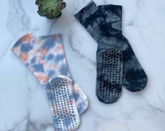 May include: Two pairs of tie-dye socks with non-slip grips. One pair features a pastel peach and blue pattern, while the other is a dark blue and gray tie-dye. A small succulent plant is visible in the background.