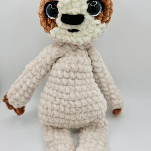 May include: A handmade crochet sloth doll with a cream-colored body and head, accented with brown patches around the eyes and on the feet and hands. The sloth has large, shiny black eyes and a black nose. The plush toy is designed to sit upright.