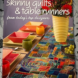 May include: A book titled "Skinny Quilts & Table Runners" edited by Eleanor Levie. The cover features a colorful table runner with spiral designs, alongside square plates, a vase of flowers, and a yellow vase.