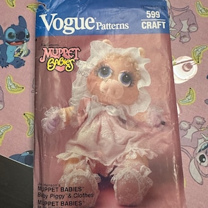 May include: A vintage Vogue Patterns craft pattern featuring a Muppet Babies Baby Piggy doll. The pattern packet is primarily pink with a blue top section. The image of Baby Piggy is wearing a pink dress and bonnet. The pattern number is 599.