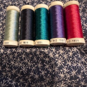 May include: Five spools of thread in various colors: light blue, navy, teal, purple, and red. Each spool has a white top with black text. The threads are arranged in a row against a patterned blue background.