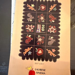 May include: A wool table rug design with a dark background and various patriotic-themed appliqués, including stars, hearts, and figures. The rug measures 26.5 inches by 41.5 inches. The design is by Laurene Sinema Designs.