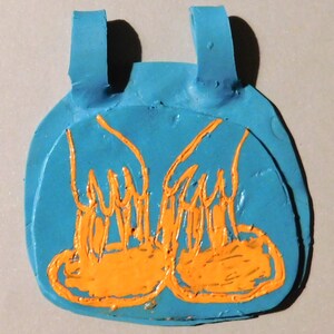May include: A handmade, blue, clay pendant with orange painted details. The pendant has a rounded shape with two straps at the top and an orange design on the front. The design appears to be a pair of feet.