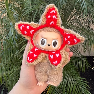May include: A plush toy with a Demogorgon-inspired crochet hat. The hat is tan with red accents and a star-like shape. The toy has a round face with large eyes and a small mouth. The body is brown and fuzzy.