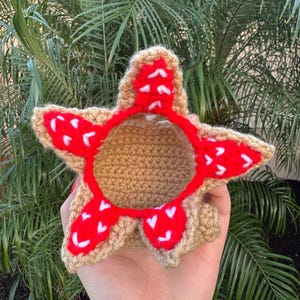 May include: A handmade crochet Demogorgon, featuring a tan body and a red, petal-like crown with white accents. The craft item is held against a backdrop of green foliage.