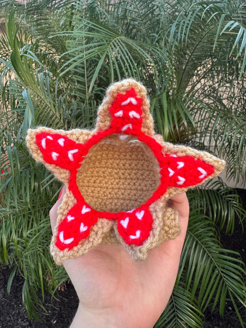May include: A handmade crochet Demogorgon, featuring a tan body and a red, petal-like crown with white accents. The craft item is held against a backdrop of green foliage.