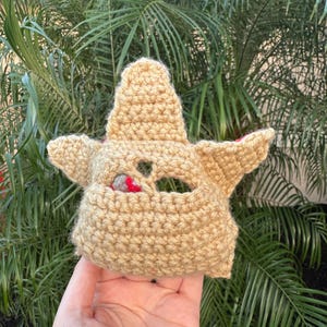May include: A tan crocheted mask with pointed ears and a small opening for the eyes. The mask has a textured appearance and is likely handmade. The background features green palm leaves.