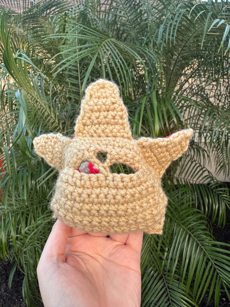 May include: A tan crocheted mask with pointed ears and a small opening for the eyes. The mask has a textured appearance and is likely handmade. The background features green palm leaves.