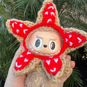 May include: A plush toy with a tan body and a star-shaped crochet hat in tan and red. The red sections of the hat have white heart-shaped accents. The toy has large, round eyes and a small, toothy grin.