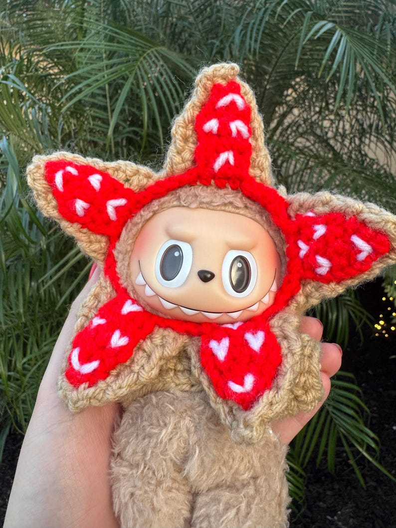May include: A plush toy with a tan body and a star-shaped crochet hat in tan and red. The red sections of the hat have white heart-shaped accents. The toy has large, round eyes and a small, toothy grin.