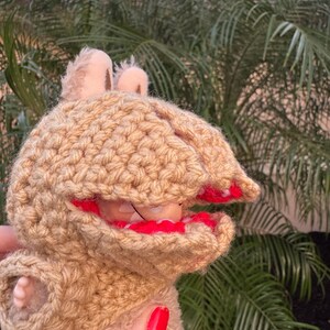 May include: A handmade crochet doll with a tan body and a large, open mouth lined with red. The doll has small, pink ears and is held against a backdrop of green foliage. The doll's mouth is open, revealing a small face.
