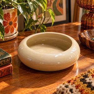 Peut inclure: Un bol en céramique rond et bas avec une finition lisse et brillante de couleur crème. Le bol présente un bord épais et arrondi ainsi qu'un bassin large et peu profond, offrant un design simple et fonctionnel avec une couleur beige uniforme.